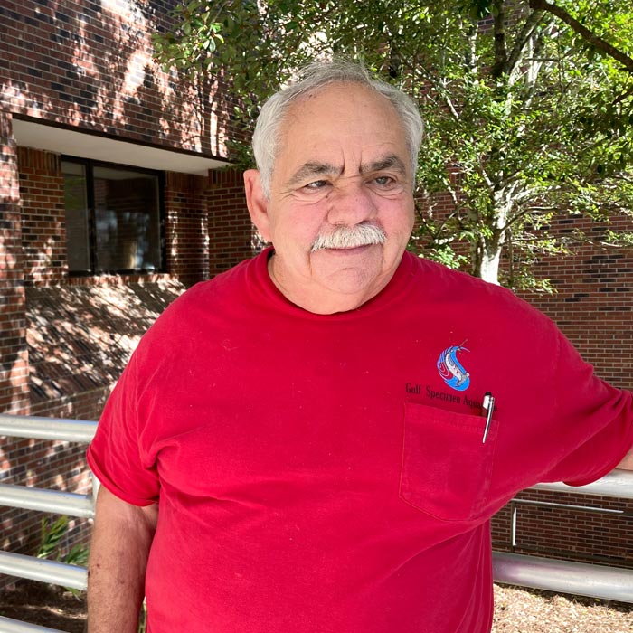 A man in a red shirt stands in front of greenery and a red-brick wall.